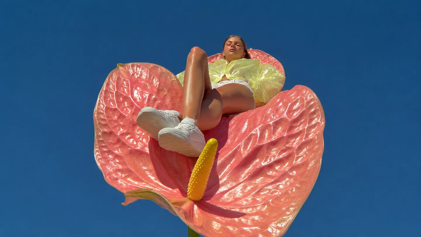 Mannequin mode assis sur une fleur rose géante sous un ciel bleu dégagé, avec un style éditorial estival en contre-plongée